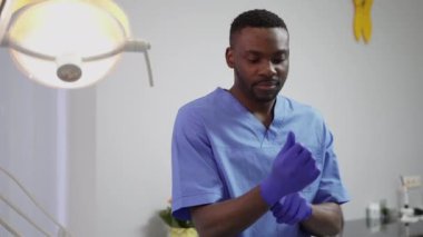 Modern dişçilik kliniği. Genç, yakışıklı Afrikalı doktor dişçi bir diş tedavi odasında duruyor ve tedavi edilmeden önce mavi lastik koruyucu eldivenler giyiyor.