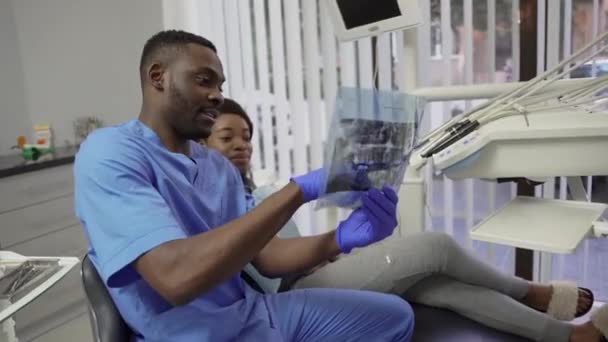 Férfi afrikai fogorvos beszél egy pácienssel, elég sötét bőrű lánnyal, a fogászati klinikán, aki körpanorámás fogászati röntgent mutat az állkapcsáról. Fogászat és fogápolás — Stock videók