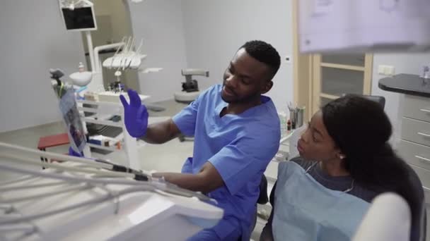 Fogászat, egészségügy, fogápolás. Fiatal jóképű afrikai férfi fogorvos megvitatja a stratégia a kezelés a beteg afrikai bájos fiatal hölgy, mutatja röntgen képet jaws — Stock videók