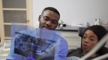 Mavi takım elbiseli ve mavi eldivenli yakışıklı Afrikalı bir dişçi, dişlerinin panoramik diş röntgenini ve dişlerinin tedavisini gösteriyor.