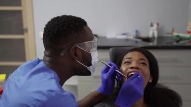 Genç Afro-Amerikalı kadın diş hekimlerinin muayenehanesini ziyaret ediyor. Ağız boşluğunun önlenmesi ve tedavisi için. Kız ve erkek doktor dişlerini kontrol ederken. Sağlıklı yaşam tarzı, sağlık ve tıp konsepti.