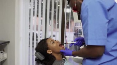 Genç Afrikalı erkek dişçi diş dolgularını özel bir ışıkla onarıp kurutuyor ve elinde polimerizasyon lambası tutuyor. Düzenli kontrol ve dolum için dişçiye giden kadın hasta