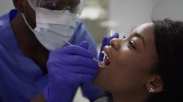 Close-up de muito africano jovem paciente sendo examinado pelo médico dentista americano africano masculino em terno azul e luvas com ferramentas odontológicas profissionais na clínica odontológica moderna — Vídeo de Stock