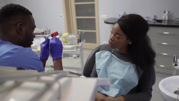 Młody, przystojny, uśmiechnięty afrykański dentysta, pokazujący model zębów swojej afrykańskiej pacjentce w klinice. Koncepcja kliniki dentystycznej, opieka stomatologiczna — Wideo stockowe
