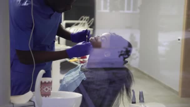 Młoda afrykańsko-amerykańska kobieta odwiedzająca gabinet dentystyczny w celu zapobiegania i leczenia jamy ustnej. Afrykański lekarz i ładna dziewczyna podczas badania zębów. Widok przez szybę okna — Wideo stockowe