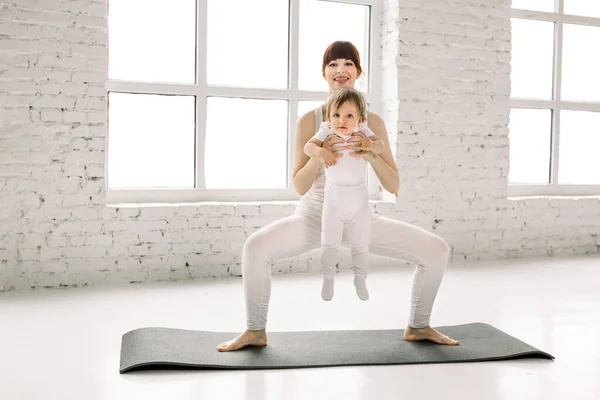Die Hubsche Junge Mutter In Weisser Sportkleidung Macht Fitnessubungen Und Hockt Mit Ihrem Kleinen Baby Auf Einer Schwarzen Matte Sport Mutterschaftskonzept Tochter Turnen Stock Photo 317076632