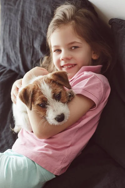 Portrait of little girl hugging with puppy of Jack Russell Terrier dog on the bad with black sheets