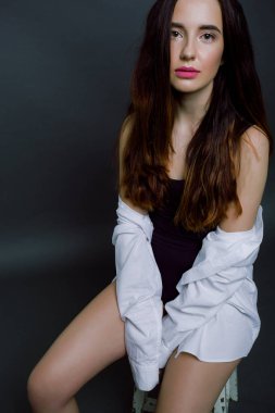 Beyaz gömlekli genç bir kadın omuzlarını açıkta bırakıyor ve bir sandalyeye oturuyor. Eller aşağı ve bacaklar arasında katlanmış..