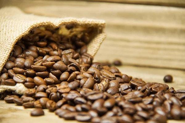A close up of a burlap bag of coffee beans spilled onto a wood table with the focus on the beans closest to the burlap bag.