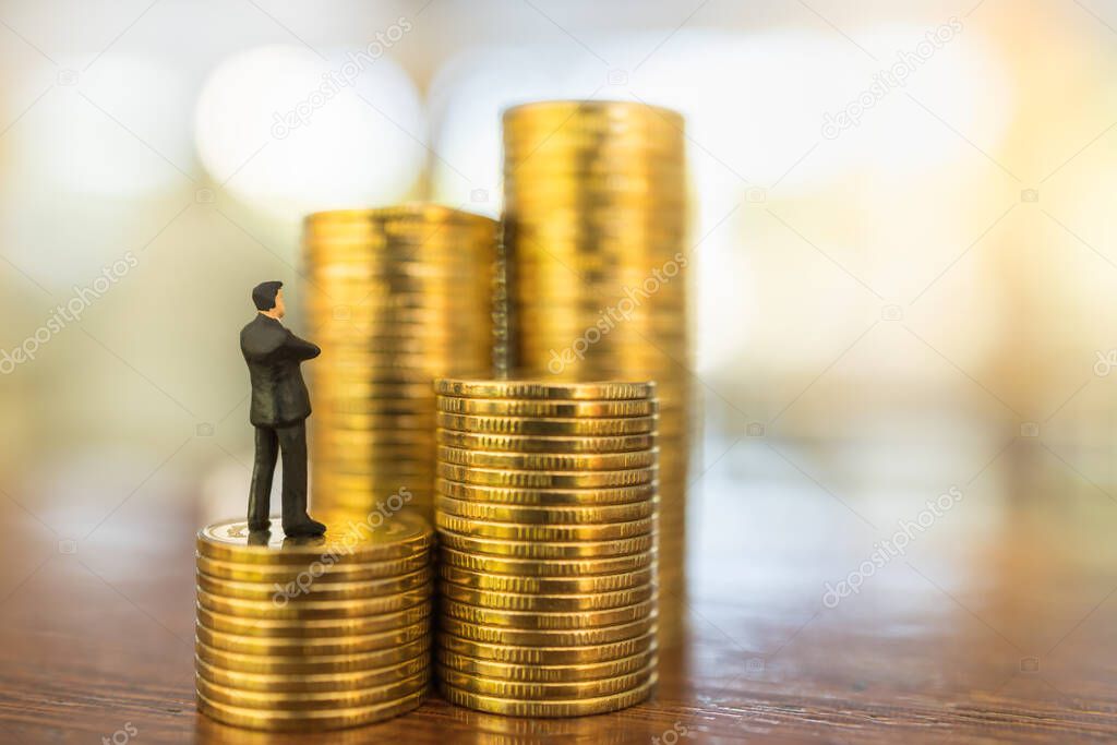 Business, Money Investment and Planning Concept.  Close up of businessman miniature people figure looking and standing on stack of gold coins on wooden table with copy sapce.