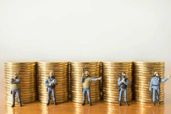 Business, Money and Security Concept. Close up of group of soldier or police miniature figure people standing and guard and protect infront of stack of gold coins on wooden table with copy space.