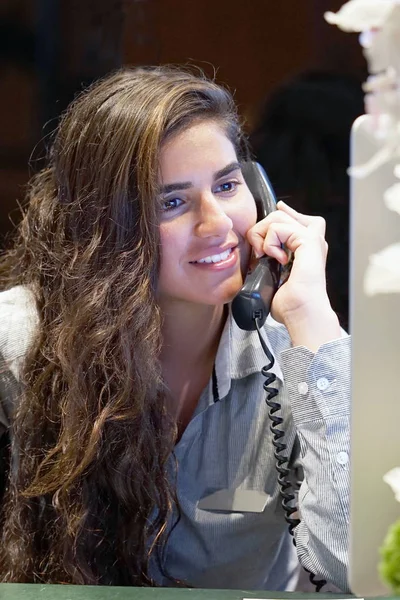 Jovem mulher sentada em sua mesa trabalhando e respondendo a um telefone ca — Fotografia de Stock