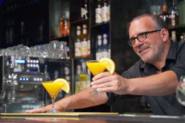 The bartender with eyeglasses wipes out the wine glass at work. Evening time. Fresh cocktail with orange.