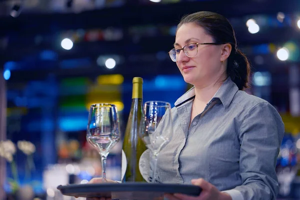 The waiter serves a table. Waitress carries a wine glasses and bottle wine on a tray in hotel restaurant, bar. The concept of service.
