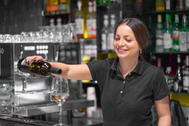 Barmen kadın gülümsüyor, beyaz şarabı şişeden bardağa dolduruyor. Arkasında alkol şişeleri olan raflar..