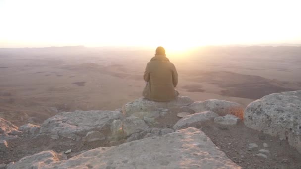 El hombre sentado en una roca en el borde de un acantilado, el sol está saliendo en el fondo — Vídeos de Stock