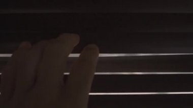 Karanlık bir odada, bir el penceredeki bir çatlağı açar ve dışarıya bakmak için kör olur.