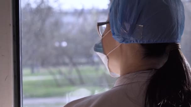 Mengukur terhadap pencegahan transmisi. Dokter wanita muda melihat melalui jendela, memakai topeng dan kacamata dalam wabah pandemi coronavirus. Hentikan COVID-19 infeksi. ProRes 422 klip, ditembak di 4K UHD. — Stok Video