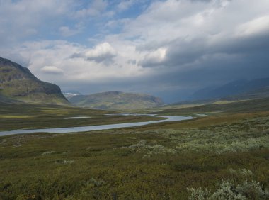 Mavi buzul nehri, huş ağacı çalıları, karlı dağlar ve dramatik bulutlarla dolu güzel Laponya tabiatı. Kuzey İsveç Yazı Kungsleden yürüyüş parkurunda.