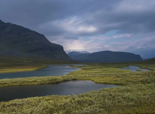 Vackert vildmarkslandskap med blå glacialflod, björkbuskar, snötäckta berg och dramatiska moln. Norra Sveriges sommar på Kungsledens vandringsled. — Stockfoto