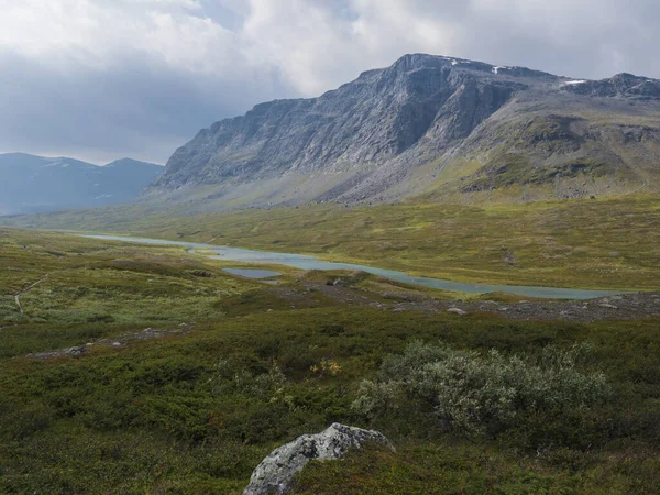 Vackert vildmarkslandskap med blå glacialflod, björkbuskar, snötäckta berg och dramatiska moln. Norra Sveriges sommar på Kungsledens vandringsled. — Stockfoto