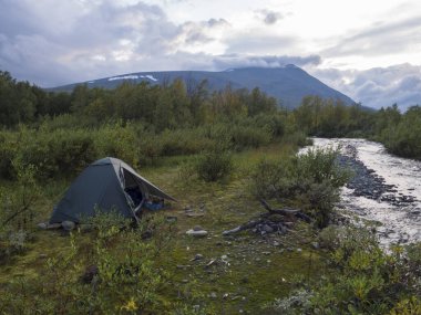 Mavi nehri, huş ağaçlarını ve dağları olan güzel Laponya doğa manzarasında küçük yeşil bir çadır. Kuzey İsveç yazı Kungsleden yürüyüş parkurunda. Mavi gökyüzü dramatik bulutlar