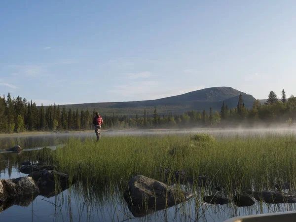 Pescador hombre la captura de peces en el lago Sjabatjakjaure en Hermosa niebla de la mañana soleada niebla en Suecia Laponia naturaleza. Montañas, abedules, bosques de abetos. Cielo azul —  Fotos de Stock