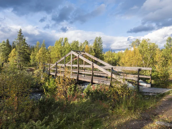 Fa láb híd folyó felett patak közelében Kvikkjokk Svédországban Lappföld nyírfa és lucfenyő erdő. Kora őszi színek, napfény, kék ég fehér felhők. — Stock Fotó