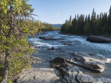 İsveç Laponya 'nın Kvikkjokk kentindeki mavi Kamajokk nehri, dağlar, kayalar ve ladin ağacı ormanıyla güzel kuzey manzarasında balık avlayan balıkçı. Yaz güneşli bir gün, beyaz bulutlar