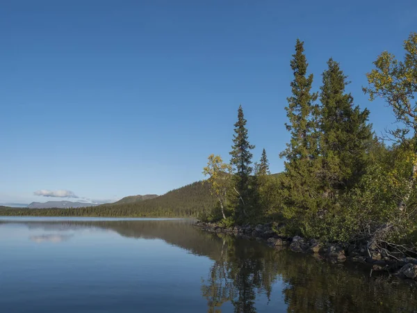 美丽的早晨在瑞典拉普兰的Sjabatjakjaure湖上。 山岭，桦树，云杉林，岩石巨石和草地。 天空，云彩和清澈的水. — 图库照片