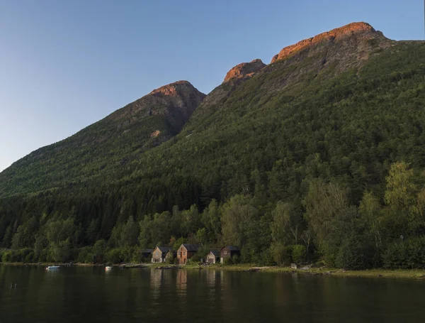 Вид на традиційний дерев'яний будиночок уздовж фіорду небезпеки в Кінсарвіку в Норвегії з лісовим скелястим пагорбом, рибальськими човнами і відображенням у бухті. Норвезький пейзаж, золота година світло і червоне просвітлення — стокове фото