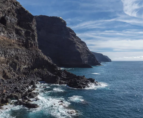 Hohe steile Vulkanfelswände aus schwarzer Lava am felsigen Ufer mit zermalmenden weißen Wellen über dem Atlantik. blauer Himmel Hintergrund. La Palma, Kanarische Inseln. — Stockfoto