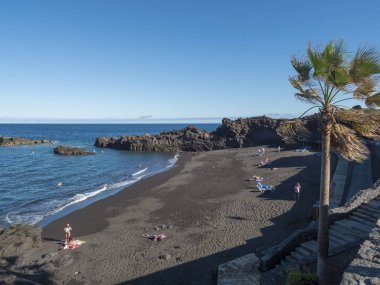 Los Cancajos, La Palma, Kanarya Adası, İspanya, 30 Aralık 2019: Güneşlenen turistlerle dolu Los Cancajos köyünün kara kumlu plajı. Lav kayalıkları ve mavi gökyüzü arka planı.