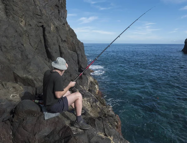 Lado posterior del joven pescador en sombrero sentado en el borde del acantilado de roca de lava con caña de pescar en la costa del océano atlántico en la isla de La Palma, España —  Fotos de Stock