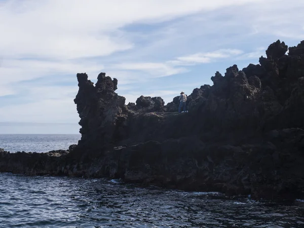 Back side of older man, local fisherman in hat standing on the edge of lava rock cliff with fishing rod at coast of atlantic ocean at La Palma island, Spain