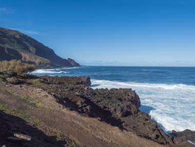 Kayalık sahildeki siyah lav volkanik kaya kayalıkları Atlantik Okyanusu üzerinde beyaz dalgaları eziyor. Mavi gökyüzü arka planı. La Palma, Kanarya Adaları.
