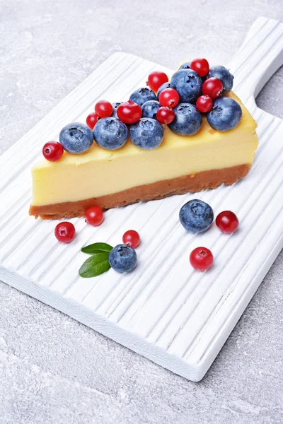 Fatia close-up de delicioso cheesecake caseiro com mirtilos frescos e cranberries na placa de corte de madeira branca — Fotografia de Stock