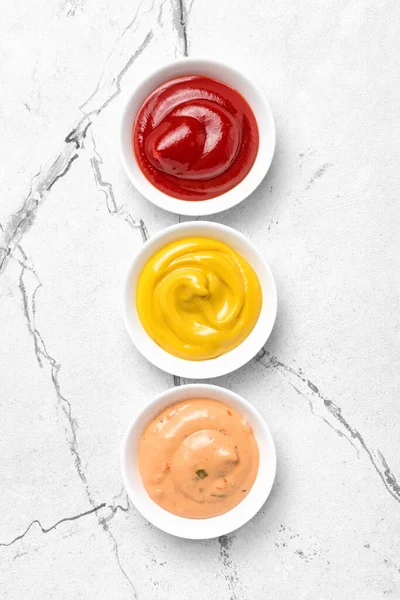 Sarı hardal, kırmızı ketçap ve kırmızı biber sosu beyaz mermer arka planda küçük kaseler halinde.