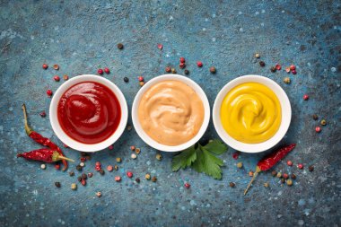 Sarı hardal, kırmızı ketçap ve kırmızı biber sosu küçük kaselerde mavi beton arka planda.