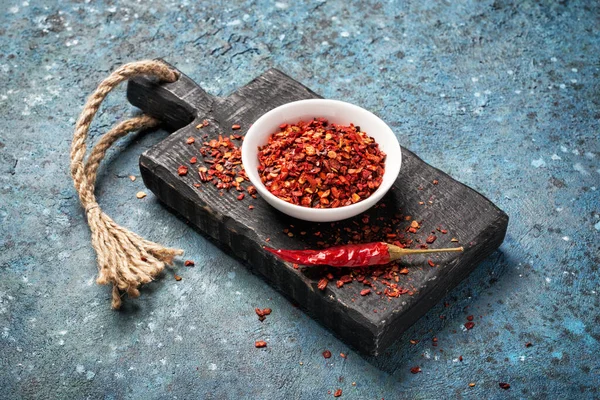 Flakes of red hot pepper flakes in white bowl and chilli pod on black wooden board and blue concrete background