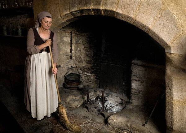 Woman dressed in renaissance maid costume using her broom to clean the fireplace in the kitchen of a property released authentic medieval castle in France