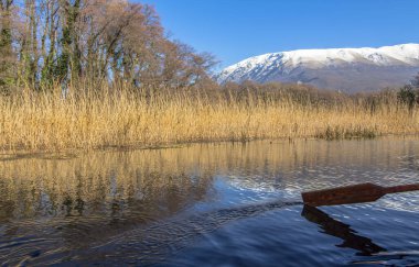 The incredible colors of the springs that flow into Ohrid Lake. Against the background of reeds, trees and snowy hills on the mountain. With a boat paddle. Ohrid Lake, Northern Macedonia.