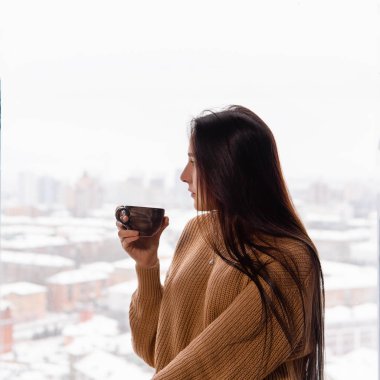 Genç bir kadın, elinde bir fincan kahveyle üst taraftaki pencerenin önünde.