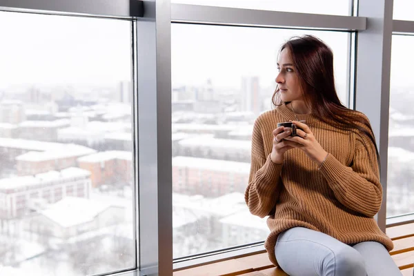 Mujer joven con una taza de café se sienta junto a la ventana en la parte superior f — Foto de Stock