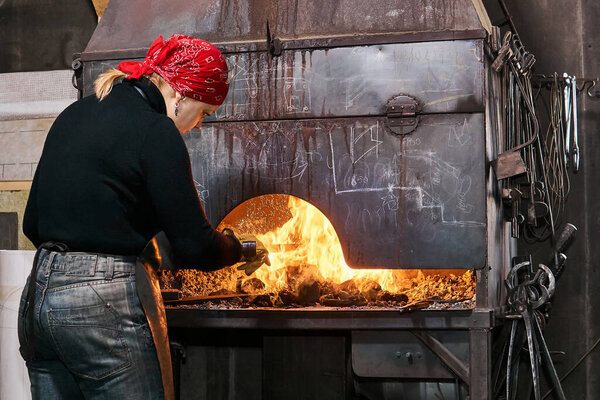 female metal artist is heating a workpiece in a blazing forge furnace