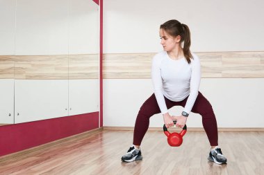 Genç bayan geniş bir spor salonunda kettlebell ile antrenman yapıyor. Aynaya bakıyor.