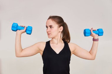 Genç, minyon bir kadın halterlerle bir egzersiz yapıyor. Yan tarafa bakıyor. Hafif bir arkaplanda yarım boy bir portre.