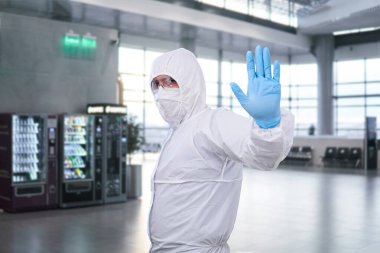 Tek kullanımlık antibakteriyel izolasyon giysisi giyen erkek epidemiyoloji uzmanı havaalanındaki ya da tren istasyonundaki bekleme odasına karşı yasaklama hareketi gösteriyor.