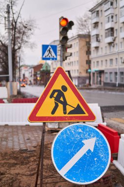 Yol tabelaları ileride ve çitin bu tarafında kaldırım işçilerinin olduğu bir caddenin önünde.