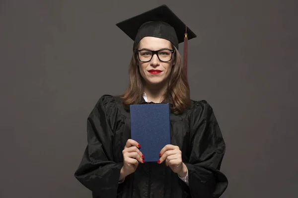 Urocza studentka nosząca czarny płaszcz i stojąca z dyplomem w rękach — Zdjęcie stockowe